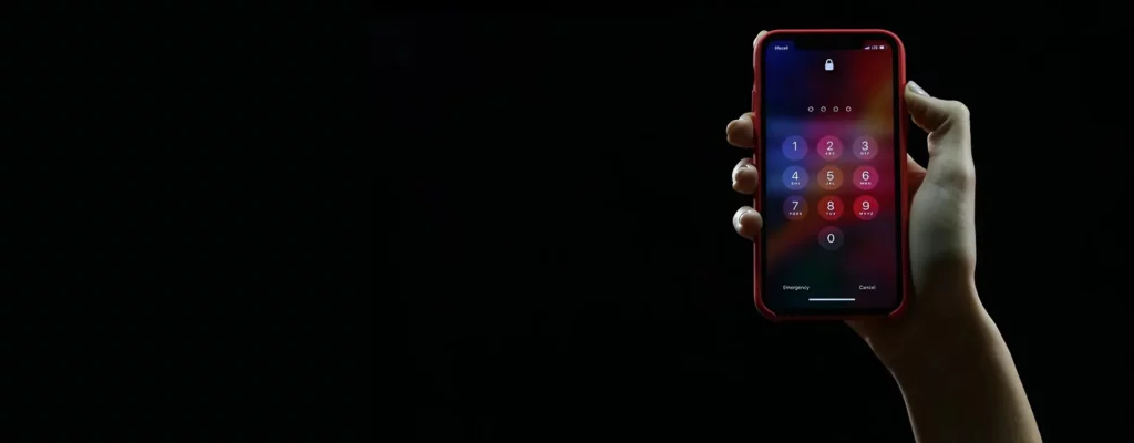 Hand holding a smartphone with the screen displaying a lock screen keypad against a dark background.