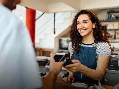 Smiling young woman in an apron accepting a payment on a mobile device from a customer at a cozy cafe.