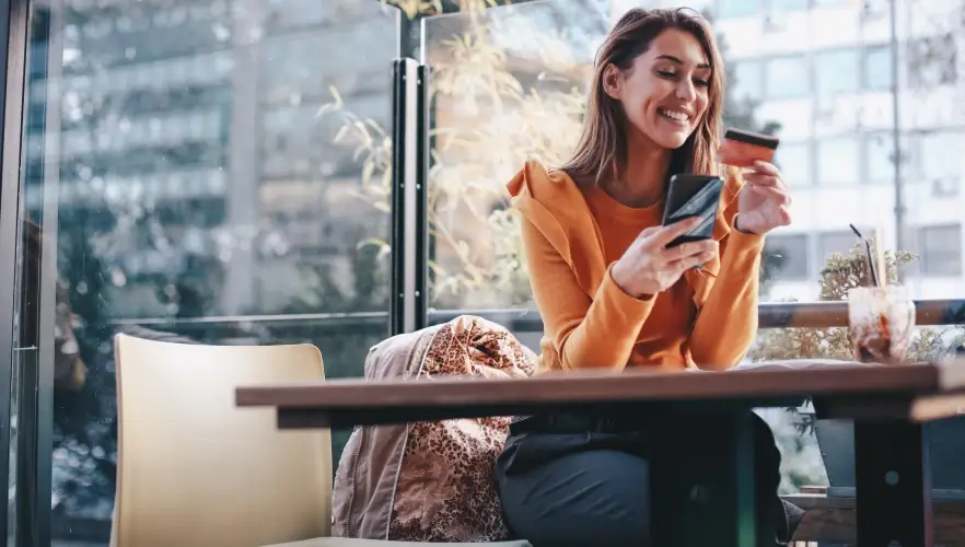 A woman enjoying a warm beverage while smiling at her phone in a cozy café setting with natural light streaming through the window.
