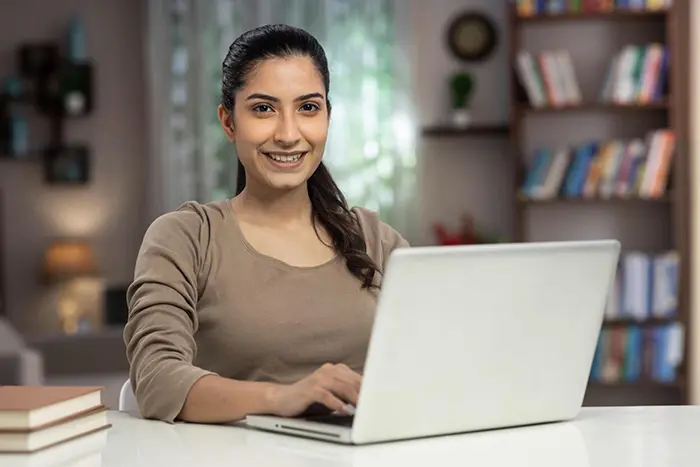 A confident woman smiling at the camera while working on her laptop in a well-lit room with bookshelves in the background.