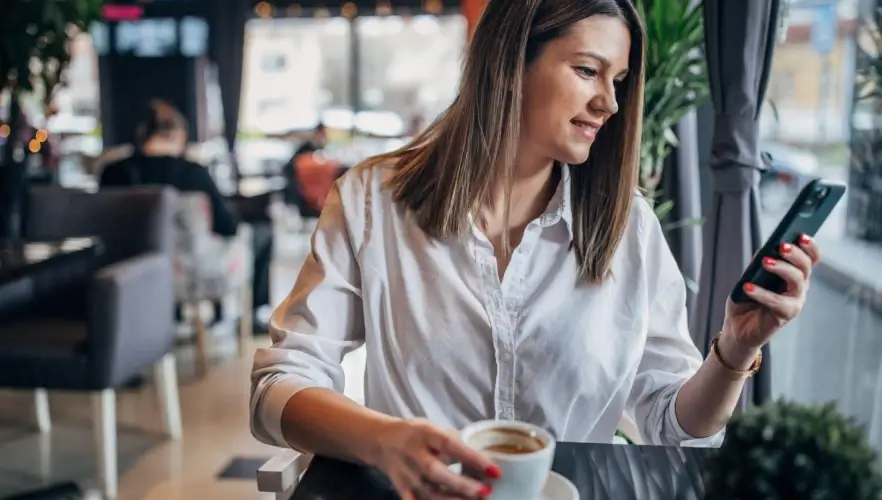 A woman in a white shirt enjoying a cup of coffee while checking her smartphone in a cafe.