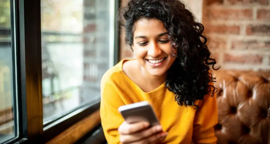 A joyful young woman in a yellow top smiling as she looks at her smartphone, comfortably seated next to a window.
