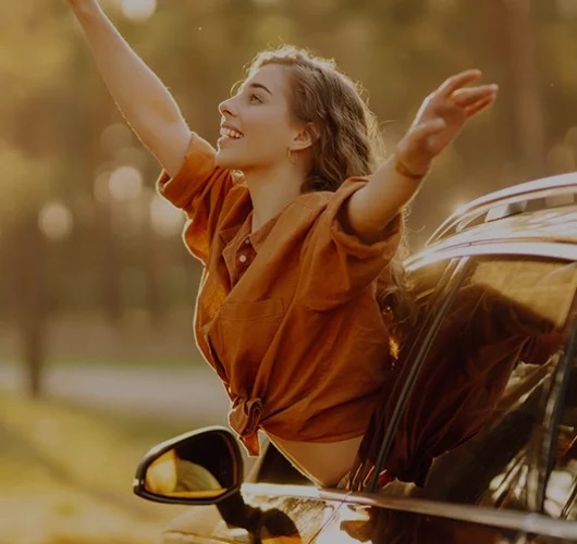 A woman joyfully leans out of a car window, arms outstretched. She wears an orange shirt, and the background features a sunlit forest, conveying freedom and happiness.
