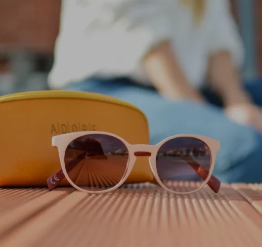 Rosegold colour sunglasses with dark lenses and an ‘Alplore’ case on a wooden surface with a reflection of a serene sky