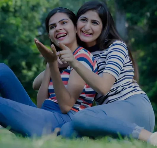 Two women, sitting on grass in a park, smile joyfully. Both wear striped shirts and jeans. Background is lush and green, creating a cheerful, relaxed tone.