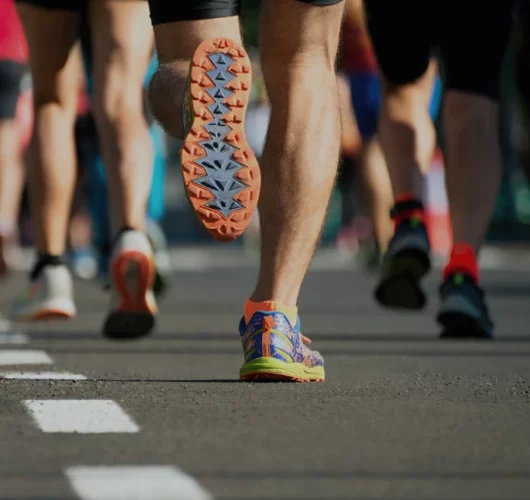 A group of runners in colorful athletic shoes is shown from behind on a sunlit road. The focus is on one runner's shoes, capturing motion and energy.