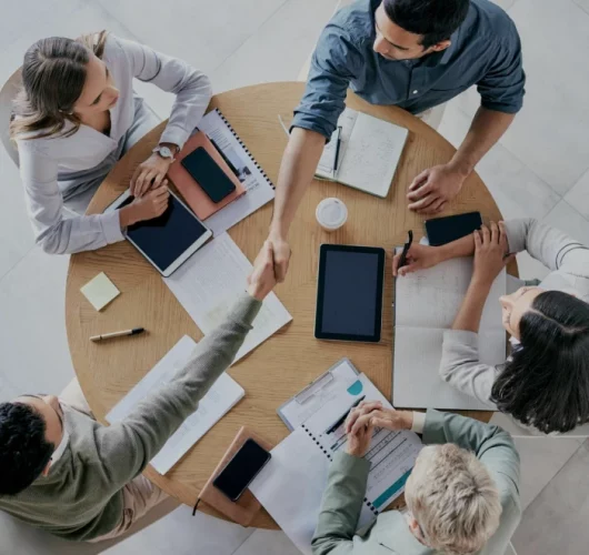 Aerial view of five people in a meeting around a round table. Two are shaking hands, while others hold tablets and notepads. The scene conveys teamwork.