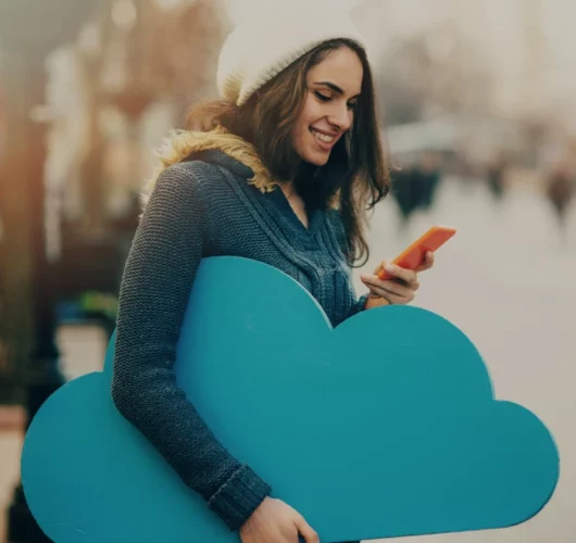A woman in a white beanie and blue coat smiles at her phone while holding a large blue cloud cutout. The background is an urban street scene.