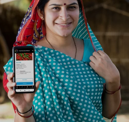 A smiling woman in a blue polka-dotted sari shows a smartphone displaying a food security app with red chili details. Her expression is proud and confident.