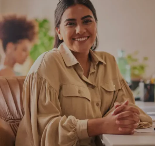 Smiling woman in a beige blouse sits at a desk with hands clasped. Blurred background shows a person and green plants, conveying a warm, inviting atmosphere.