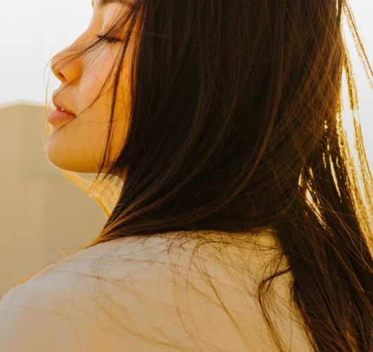 A woman with long, dark hair is softly backlit by the sun. Her eyes are closed, and she wears a peaceful expression, with hair gently blowing in the breeze.
