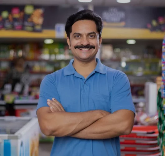 financial-empowerment Smiling man with a mustache in a blue shirt stands confidently with crossed arms in a colorful grocery store, conveying friendliness and warmth.