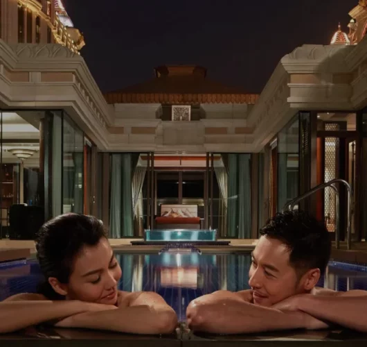 A woman and a man relax in a luxurious pool at night, leaning on the pool edge and smiling at each other. Elegant buildings with warm lights are visible in the background.