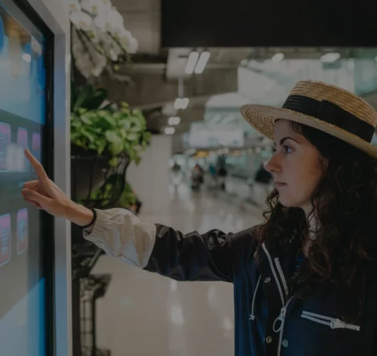 A woman in a hat uses a touchscreen kiosk in a modern indoor space, with plants nearby. She appears focused, and the setting conveys a tech-savvy, contemporary atmosphere.