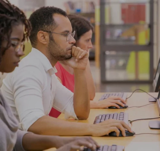 Three focused individuals sit side by side at computers in a library, with an atmosphere of concentration and quiet determination.
