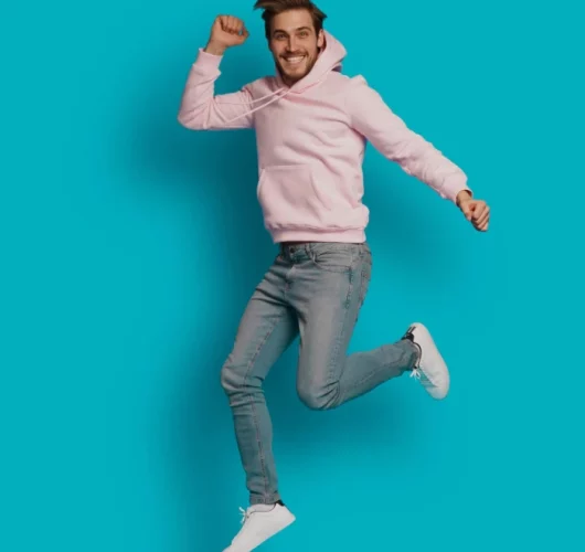 A person in a pink hoodie and jeans joyfully jumps against a bright blue background, conveying happiness and energy.
