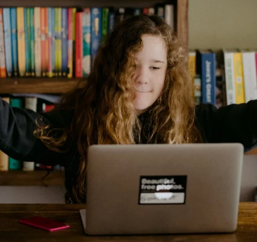 A young girl with curly hair raises her arms in excitement while looking at a laptop. Bookshelves are in the background, creating a studious atmosphere.