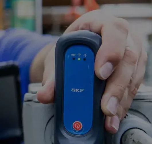 A hand holds a blue SKF diagnostic tool over machinery, capturing data. The environment is industrial, suggesting focus on equipment maintenance.