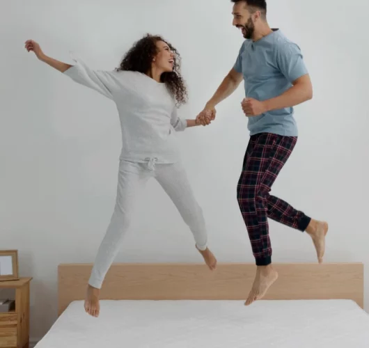 A joyful couple jumps on a bed, holding hands. The woman wears gray pajamas, and the man wears plaid pants and a blue T-shirt, both smiling in a minimalist bedroom.
