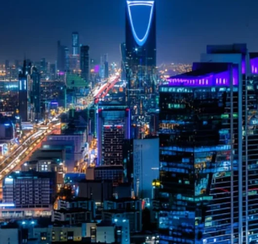 Vibrant cityscape at night featuring modern skyscrapers with bright neon lights, prominent building with distinct curved top, and busy streets below.